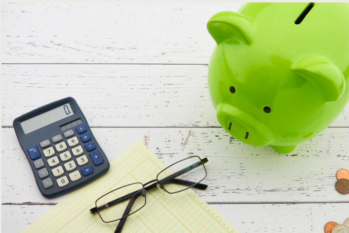 A piggy-bank, calculator, change, and glasses are laid on a white top.